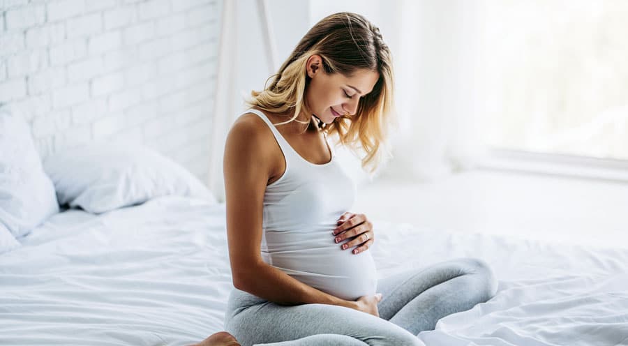 Pregnant woman sitting on her bed with hands on her belly smiling